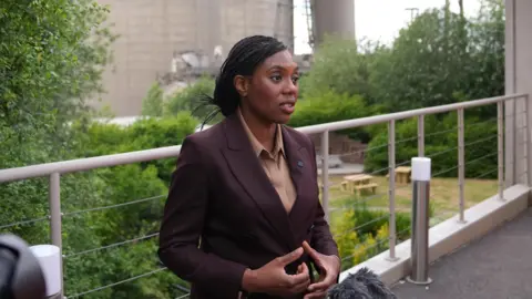 The Conservative leader Kemi Badenoch stands in front of a silo at the Cemex cement plant in Rugby. She is wearing a plum-coloured suit over an light brown blouse.