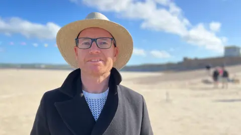 Stuart McGarry stood on the beach. He is wearing a dark coat and is wearing a sunhat.