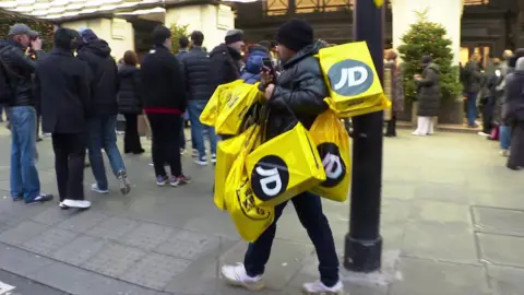 EVN / BBC A man with several shopping bags from JD sports walking past a Boxing Day queue in central London's shopping district.