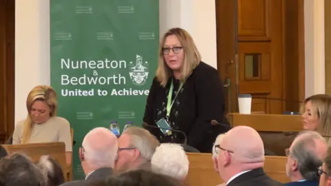 Brady Hughes stands in the chamber of Nuneaton and Bedworth Borough Council. She has blonde shoulder length hair and is wearing a dark cardigan and glasses. She is surrounded by councillors who are seated and some have their backs to the camera.