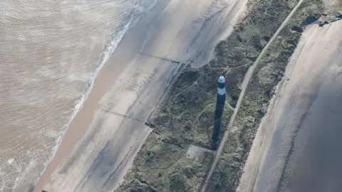 A drone view of a black-and-white lighthouse on a narrow spit of land with green dunes in the centre and sandy beaches and sea water to either side.