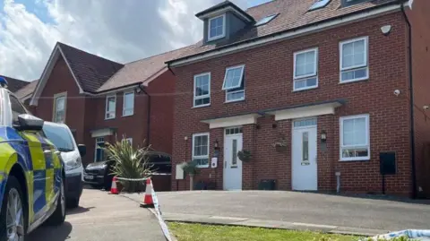 Two-storey demi-detached houses, with dormer windows in the roof. There are four white-framed windows on the first floor and two on the ground floor. There are identical white doors on both houses. There is police tape across the block-paved driveway and a white van and a police car parked outside.  