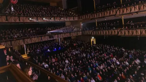 David Betteridge A packed-out Bristol Beacon main hall is seen full of audience members watching the gala evening at the Slapstick Festival. The auditorium is largely in darkness with a projector beam coming from high up at the back.