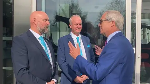 Eddie McDonald with very short hair and grey beard, Martin Griffiths with short white hair and Nigel Farage with short white hair. They are all wearing blue jackets and light blue ties. Farage has both hands up as he is explaining something. There are glass doors behind them.