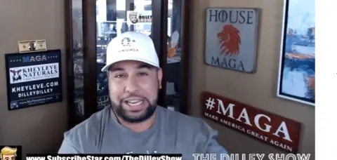The Dilley Show Brenden Dilley seen in one of his online videos, wearing a white baseball cap and staring at the camera with a big "MAGA" sign on the wall next to him 