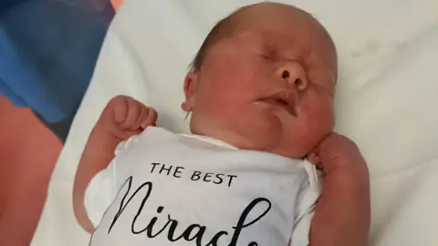 Emma Brown Newborn baby sleeps with her fists curled. Her baby grow reads 'The best miracle'.