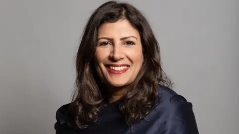 UK Parliament A woman is smiling for the camera in a portrait. She is wearing a blue top in front of a grey background.