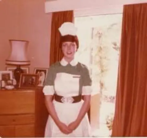 King’s College Hospital NHS Foundation Trust An old photograph of Ann Gibbs when she was younger. She is wearing traditional nursing uniform, consisting of a fitted green dress with a white collar and cuffs, a white apron, and a white nurse's cap. She is standing in front of a window with red curtains and a brown chest of drawers with a lamp and picture frames on top. 