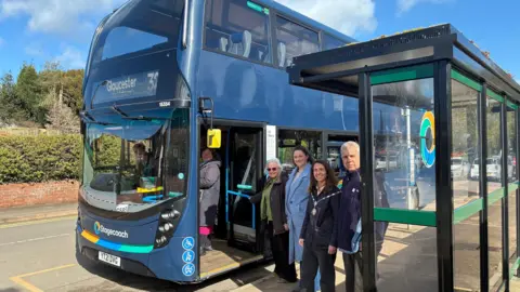 An older woman stands on a double decker bus as four other people - three women and an older man - wait by the open door at the bus stop. It is a sunny day and there is trees and bushes on the other side of the road.