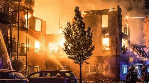 The shell of a building which is consumed by flames, is being tackled by firefighters. There are cars and a tree in the foreground. Firefighters can be seen looking up towards the building and there are jets of water being sprayed into the inferno.