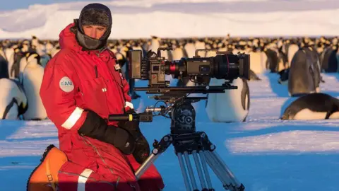 Lindsay McCrae in Antarctica kneeling on the snow with his broadcast camera on a tripod, with dozens of Emperor penguins behind him. McCrae is wearing a red snow suit, orange boots, black balaclava-style black hat and black mittens. He is looking at the camera.