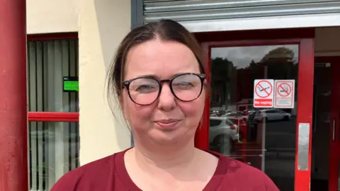 A lady with brown hair tied back. She is wearing black round-lens glasses. Behind her is red doors and a cream building.