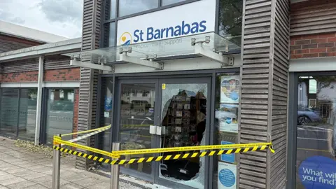 The outside of the St Barnabas shop showing the right-hand front door completely smashed there is yellow and black tape around the entrance