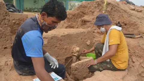 Bhakarba Thakor Abhijit Ambekar (right) who discovered the skeleton in 2019 is seen brushing soil off the skeleton, wearing a yellow t-shirt and a green hat.