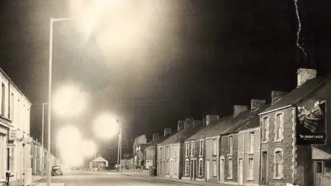 Maureen Davies/Fforestfach History Blog A black and white night-time photo of a Swansea terraced house street scene, circa 1950. A billboard for cigarettes can be see on the side of a house. It is the sort of district that Gwladys spent the latter half of her life 