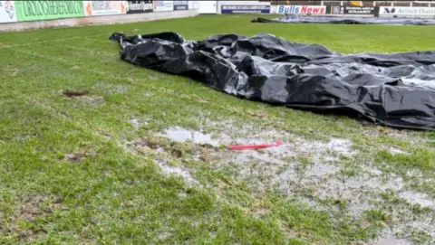 Hereford FC The pitch is holding water in small muddy/grassy puddles with a black plastic covering sat to one side.