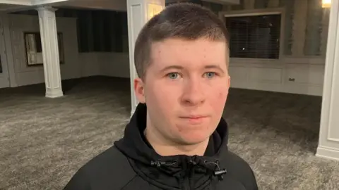 Conor looking at the camera. He has short brown hair and is wearing a black jacket.