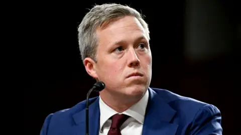 Getty Images Dan Driscoll, wearing a blue suit jacket and red tie, looks up and over to his left behind a small black microphone 