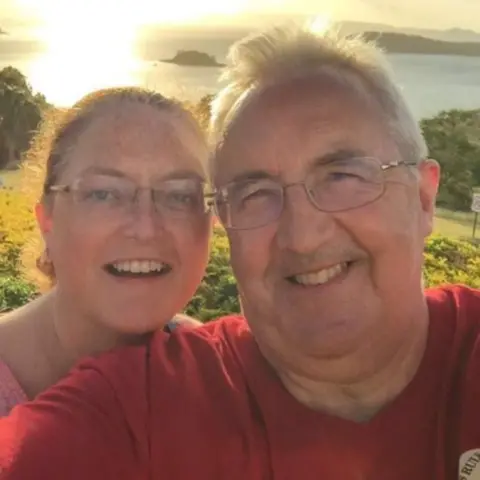 A smiling man and woman taking a close-up selfie outdoors with grassy hills and a body of water in the background.