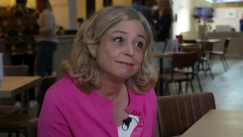 A blonde woman sitting inside. She is wearing a magenta cardigan over a white shirt.