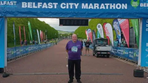 Jimmy O'Boyle Jimmy O;Boyle wears a purple t-shirt and black trousers and is standing at the finish line of the Belfast marathon