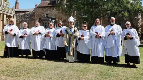 Church of England Suffolk Members of the Church of England stand together and smile at the camera on a sunny day. They wear matching white robes while a bishop in the middle wears different robes in gold, white and blue colours, and holds a staff.