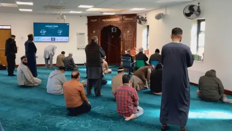Andrew Sinclair/BBC Nineteen men can be seen in a large blue-carpeted room. Some are standing, others are crouched down in prayer. 
