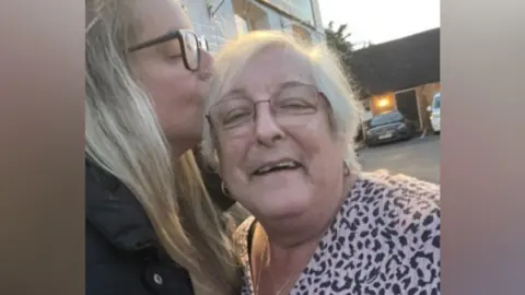 Hospice in the Weald A blonde woman, wearing glasses and a black coat, is kissing an elderly woman on the head. 
