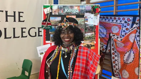 A woman in traditional Kenyan dress. A display board for Kenya is behind her.