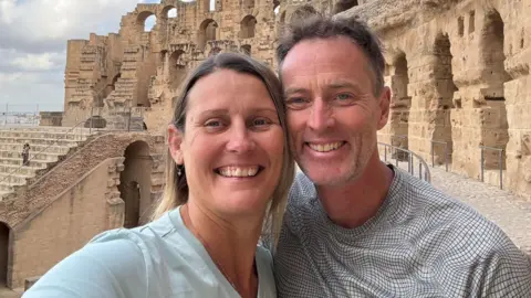 A blonde woman wearing a pale blue t-shirt is standing next to a man, with greying hair, in a checked black and white t-shirt. They are standing in front of some ancient ruins.