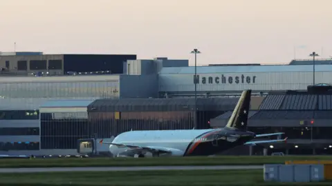 An airplane is on the runway, with grass in the foreground and an airport in the background with Manchester emblazoned on it