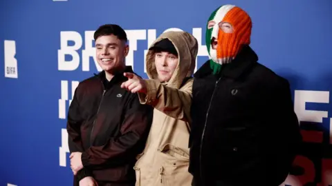 Kneecap at 2024 BIFAs - left to right Móglaí Bap in black Lacoste zip jacket, Mo Chara in a cream jacket with hood up, wearing a black snapback and pointing at camera, and DJ Próvaí in black jacket and green white and orange tricolour balaclava