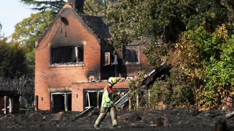 A firefighter wearing a helmet and hi-vis walks in front of a burnt out red-brick house.