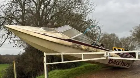 Doug McMurdo A cream coloured boat, shaped like a retro speedboat, leans against white barriers with its nose in the air, on a country lane leading into a field.
