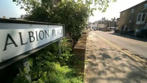 BBC A white street sign with black writing reading ALBION PLACE. The sign is on the left of the image and is surrounded by green plants. There is a pavement in front of it and houses on the right