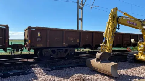 A large yellow digger parked next to a section of track carrying freight carriages 