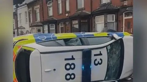 Snapchat A police car with smashed windows lies on its side in the middle of a road.