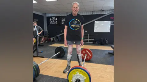 A man in his early 80s smiles as he stands in a gym between two large weights. He is wearing a black T-shirt with a large blue and gold logo, grey shorts, black trainers, and tall black socks. He has weightlifting powder all over his legs. 