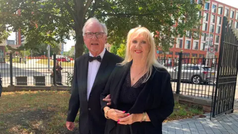 A man with grey hair and dark rimmed glasses links arms with a blonde woman in a black dress