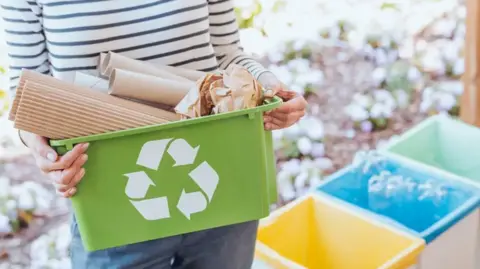 A woman in a blue stripy top holds a green recycling tub with paper in it and is stood next to other recycling bins. 