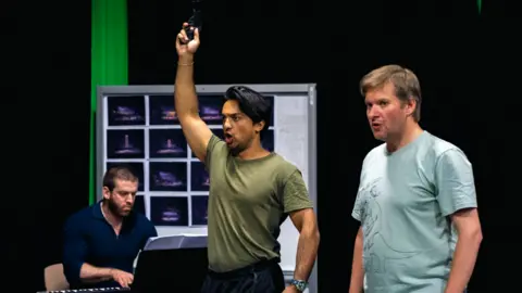 Lian Furness Three performers on stage. Two of them are standing in the foreground and one has his arm raised as he points a pistol in the air. A third person is sat down and playing a keyboard