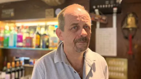 Elizabeth Baines / BBC Un hombre con un polo gris está de pie frente a un bar. Una campana se ve borrosa detrás de él.
