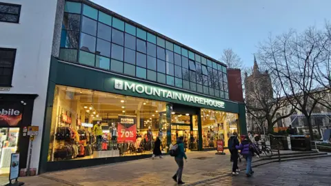 Paul Moseley/BBC A picture of the front of Mountain Warehouse. Daylight is fading so it the shop is lit up, with a number of pedestrians walking past.