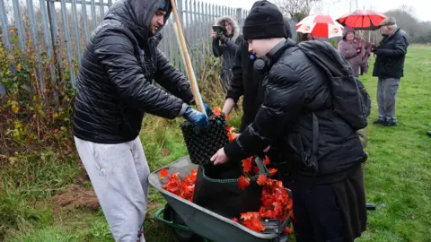 PA Media Thomas Irwin and Adam Nait from The Tree Amigos volunteer group, unload the sapling. The tree has been transported in a wheelbarrow with a garland of autumn leaves. People are standing with umbrellas behind them.
