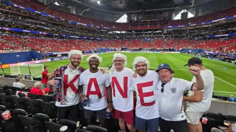 Paul Green Paul Green with 4 men wearing t-shirts that spell out KANE in red on a white background, Green has a blue bucket hat with white England shirt and behind is the football pitch.