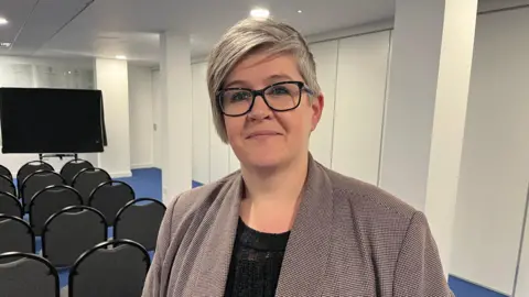 Deputy Haley Camp - A woman with grey hair, wearing black rimmed glasses. She has a grey checked jacket on over a black blouse. She is standing in front of rows of chairs, organised for a presentation. 