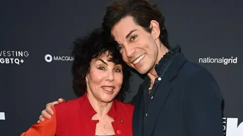 Getty Images Ruby Wax and Tom Read Wilson pose with their arms around one another. Ruby has black hair and a red jacket with a silver necklace and Tom wears a navy jacket with a blue shirt underneath with a frilled collar. Both are smiling. 