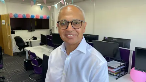A man with short grey hair and dark-rimmed glasses smiles at the camera in an office. He wears a white shirt and stands among desks with computer monitors. Pink and blue bunting and balloons are strung up behind him.