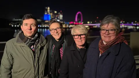 Group shot of four people in coats and scarves with a London night skyline in the distance behind them