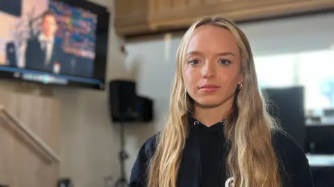 BBC Georgia has long, light-colored hair is standing indoors near a stage area. She is wearing a dark hoodie with a small white logo on the chest. In the background, there is a large screen showing another person, along with chairs and technical equipment, indicating an event or presentation setting